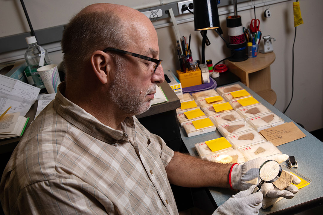 Chris Rollston examines pottery fragments with a magnifying glass while seated at a table